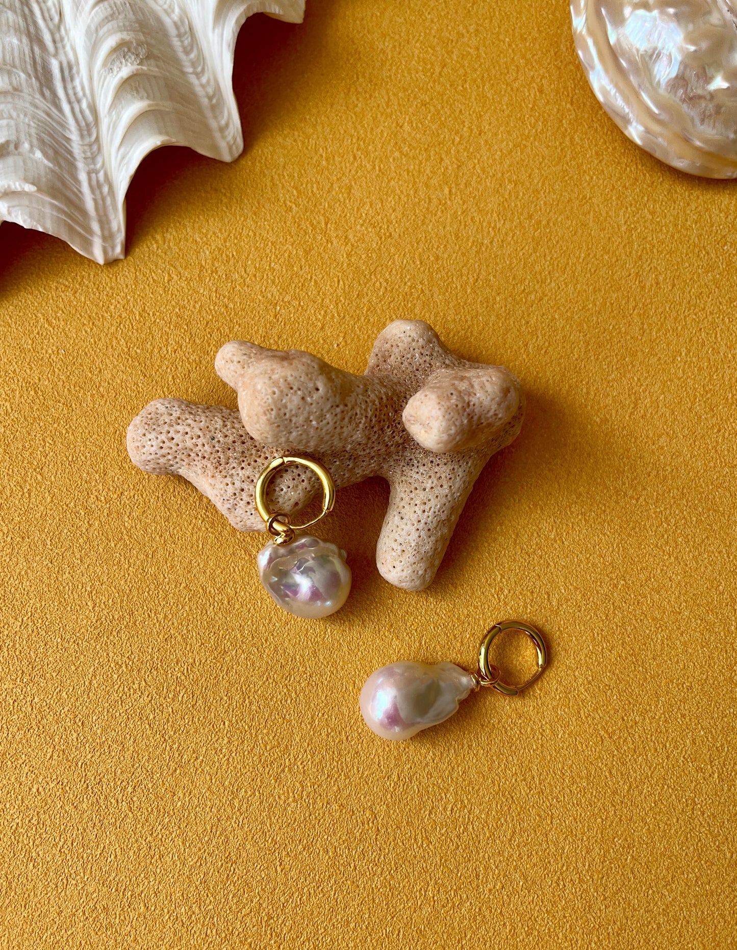 Baroque Pearl earrings with gold hooks on a textured beige surface with seashells.