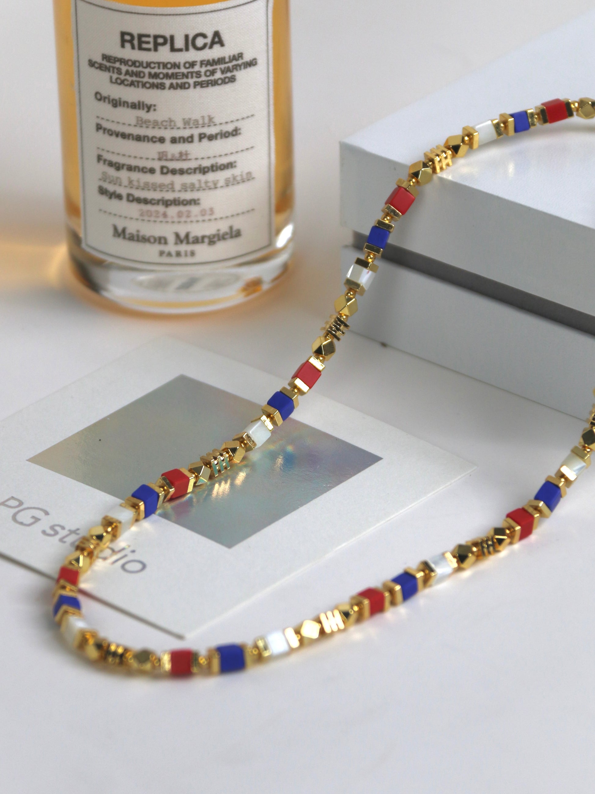 Necklace for men with gold, red, and blue beads on a white surface with a bottle labeled 'Maison Margiela' in the background.