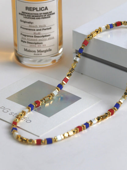 Necklace for men with gold, red, and blue beads on a white surface with a bottle labeled 'Maison Margiela' in the background.