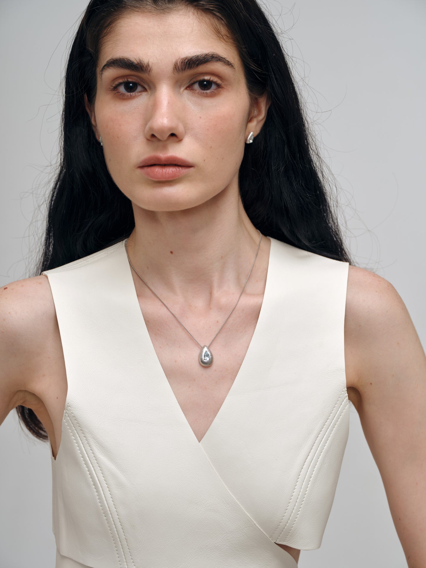 Woman wearing a white sleeveless top and a necklace with a pendant on a plain background