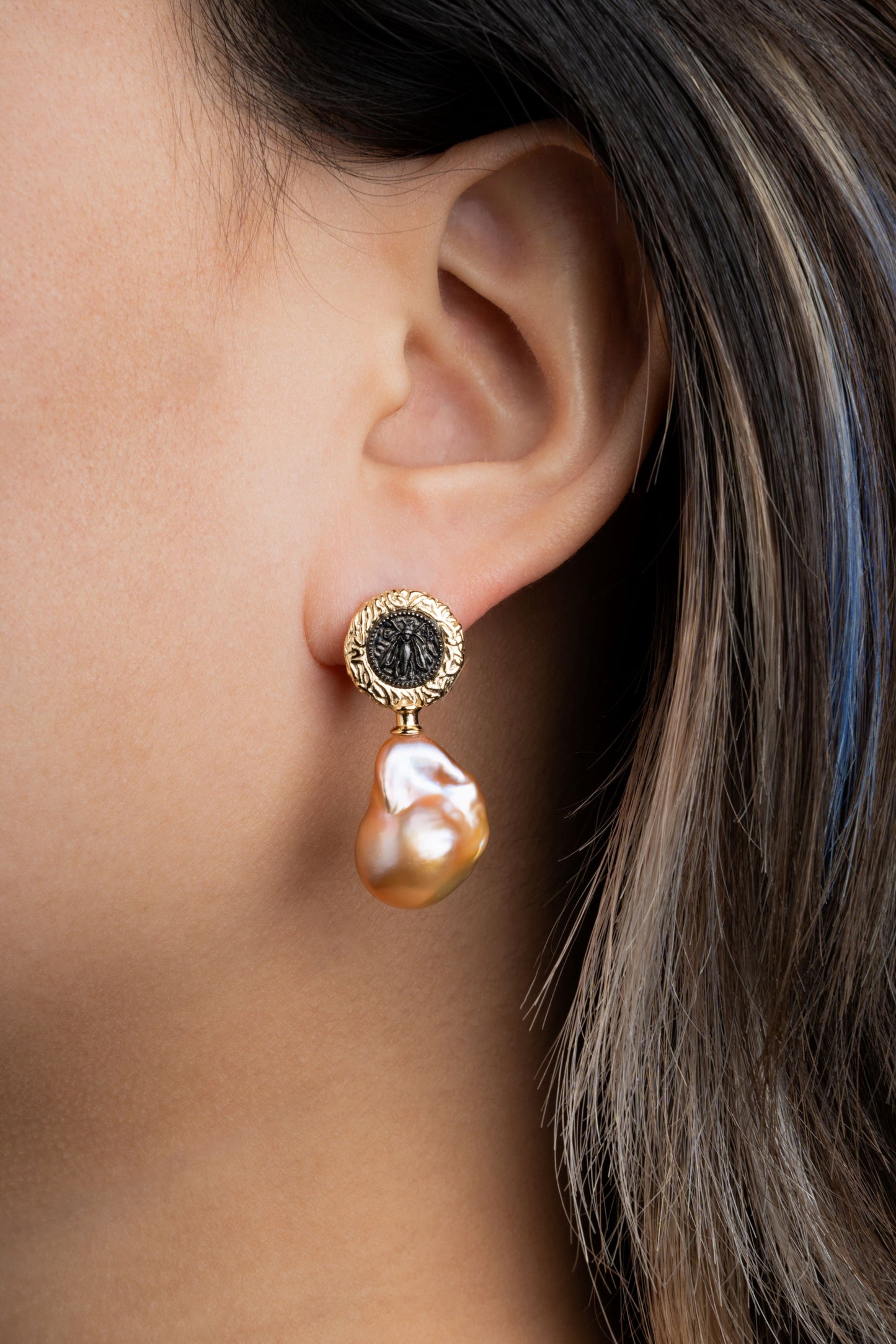 Close-up of a gold earring with a black stone and pearl drop worn by a person.