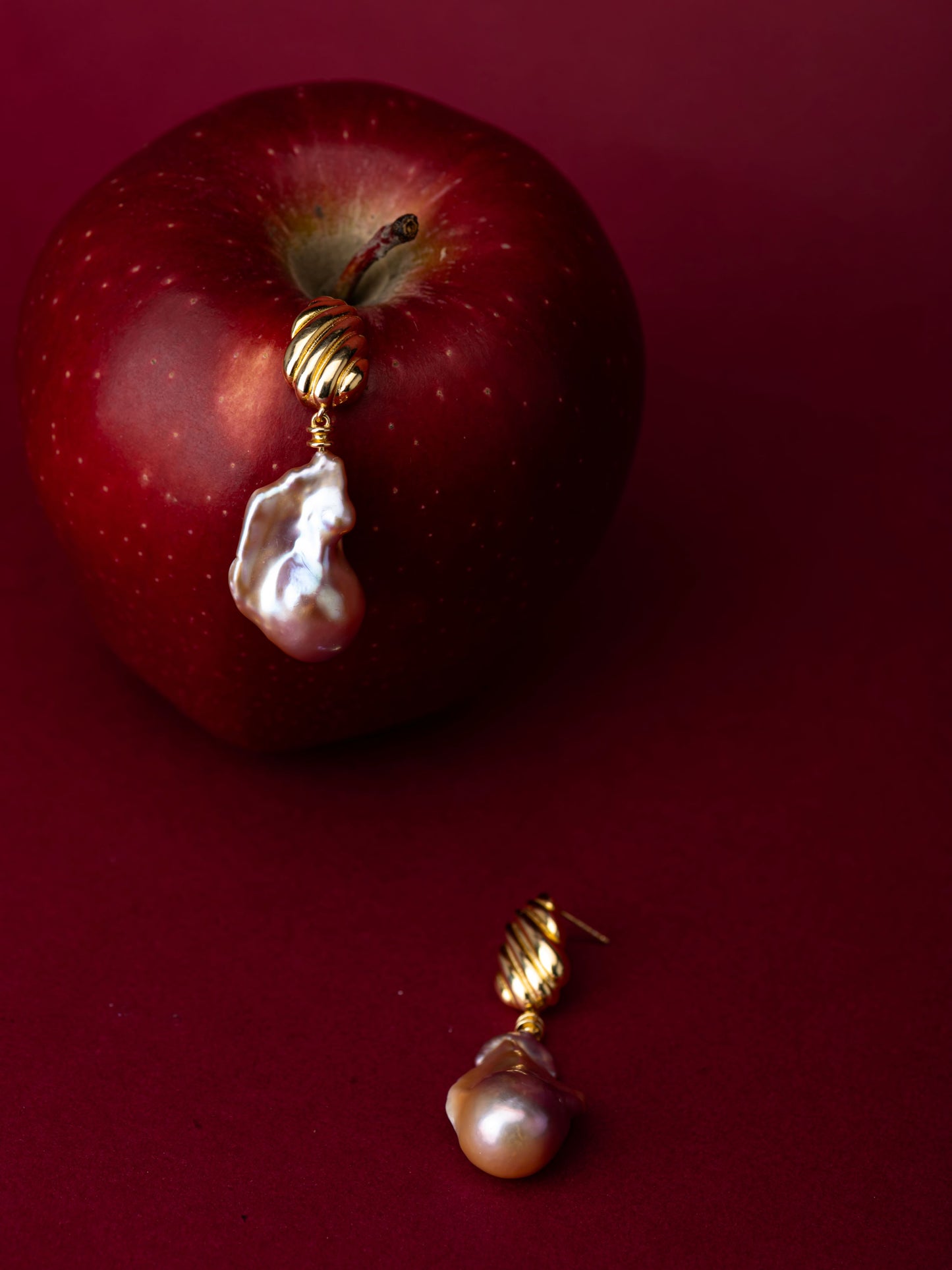 Gold earrings with a baroque pearl drop on a red apple against a red background