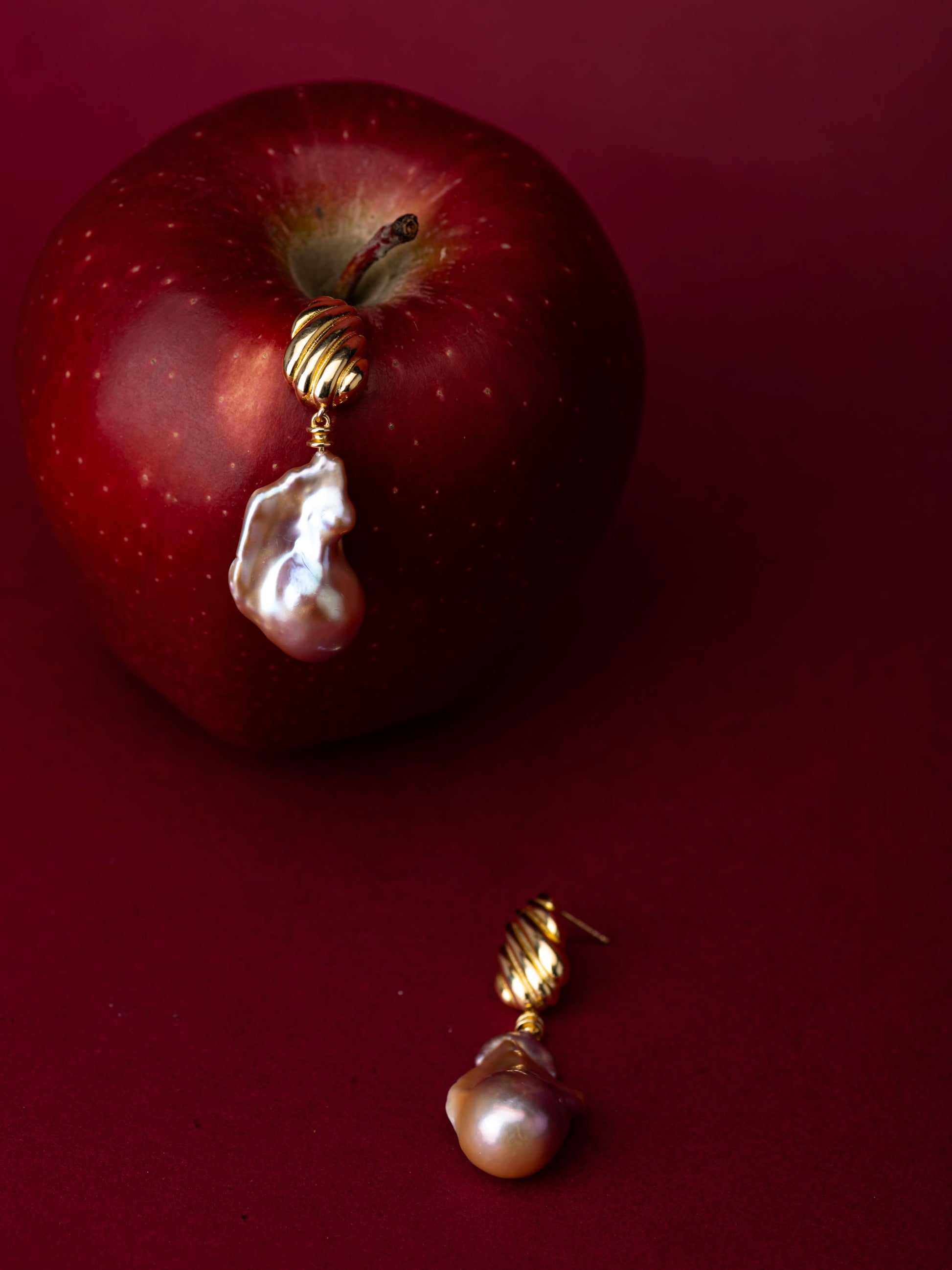 Gold earrings with a baroque pearl drop on a red apple against a red background