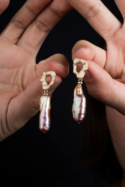Long baroque pearl earrings with high luster held between fingers against a dark background