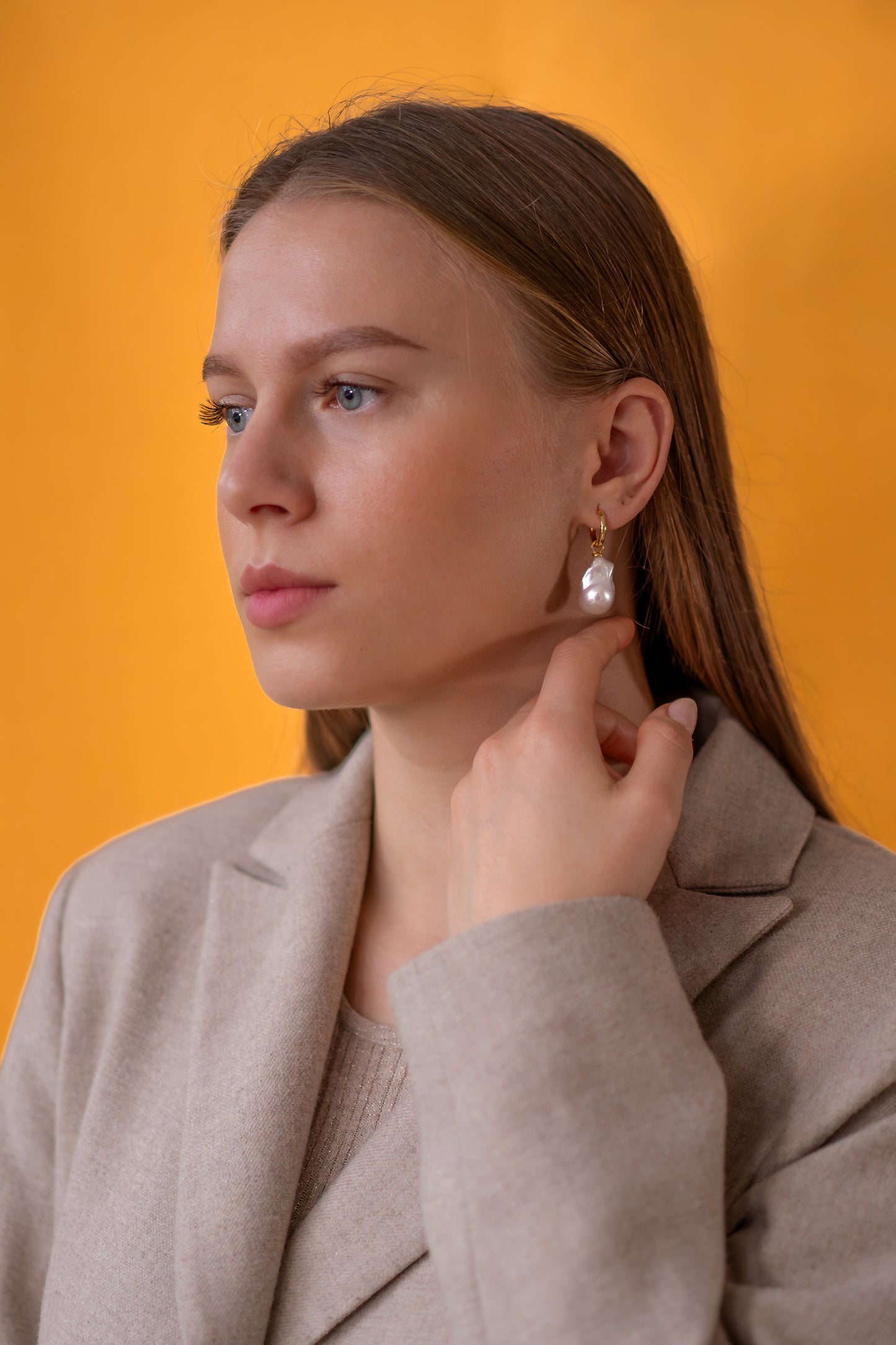 Woman wearing golden earrings with baroque pearl against an orange background