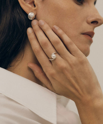 Close-up of a woman wearing pearl earrings and a ring, with a neutral background.