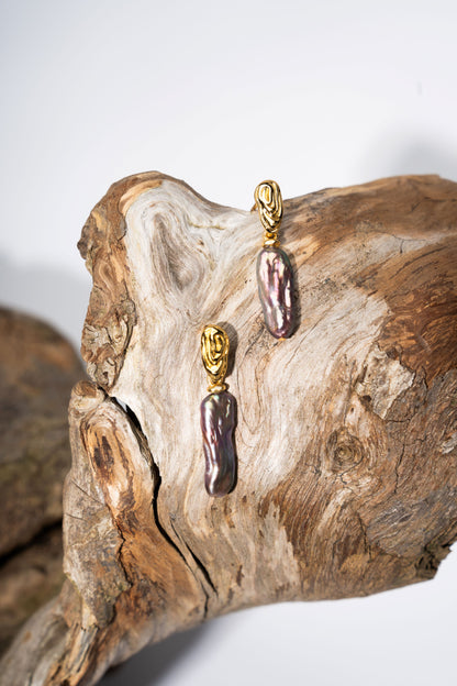 Pair of earrings with gold clasps and purple pearl on a piece of driftwood.