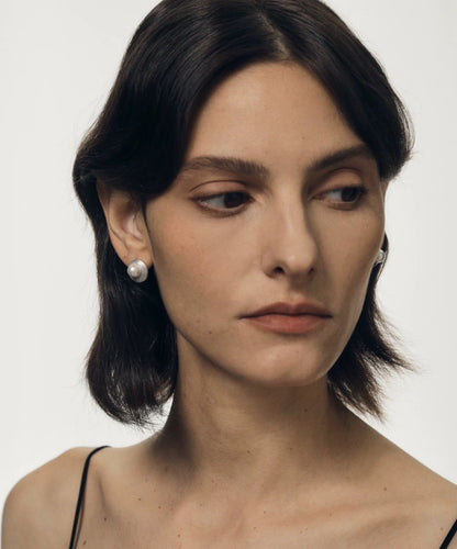 Woman with short dark hair wearing pearl earrings against a plain background