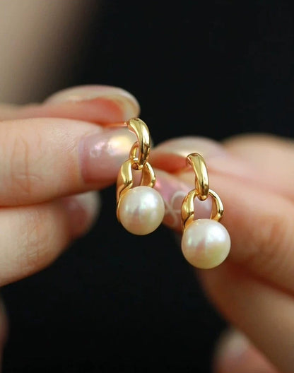 Gold hoop earrings with pearl charms held between fingers against a dark background