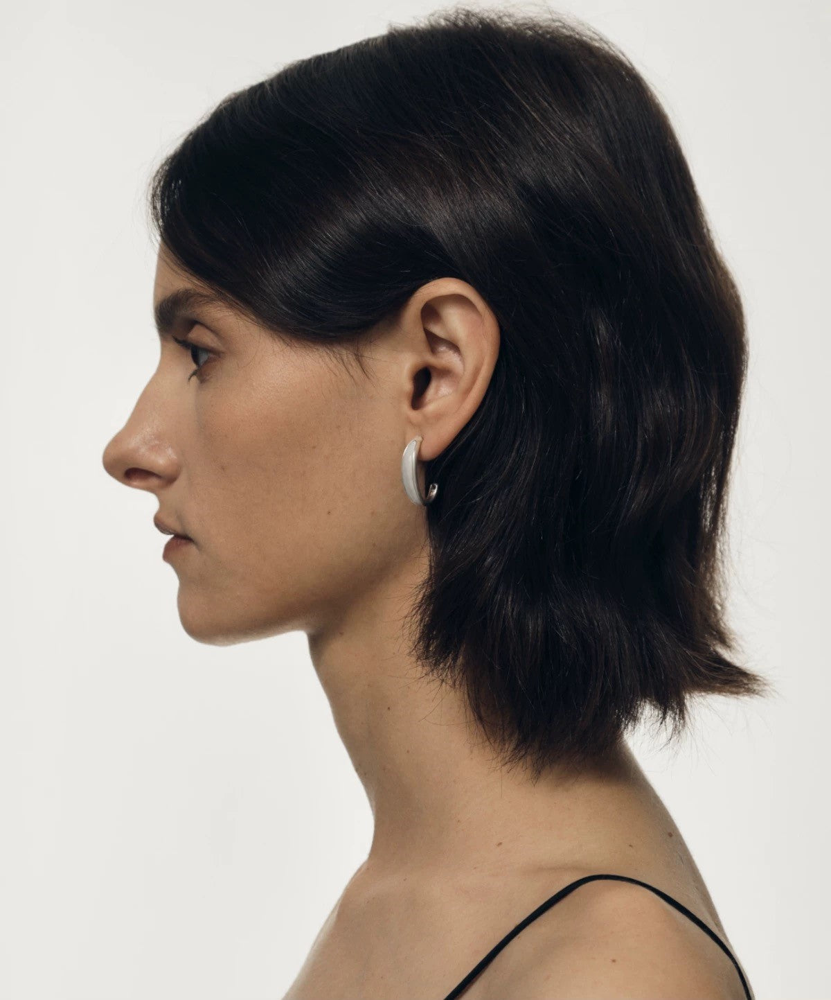 Side profile of a person with dark hair wearing a small earring on a plain background