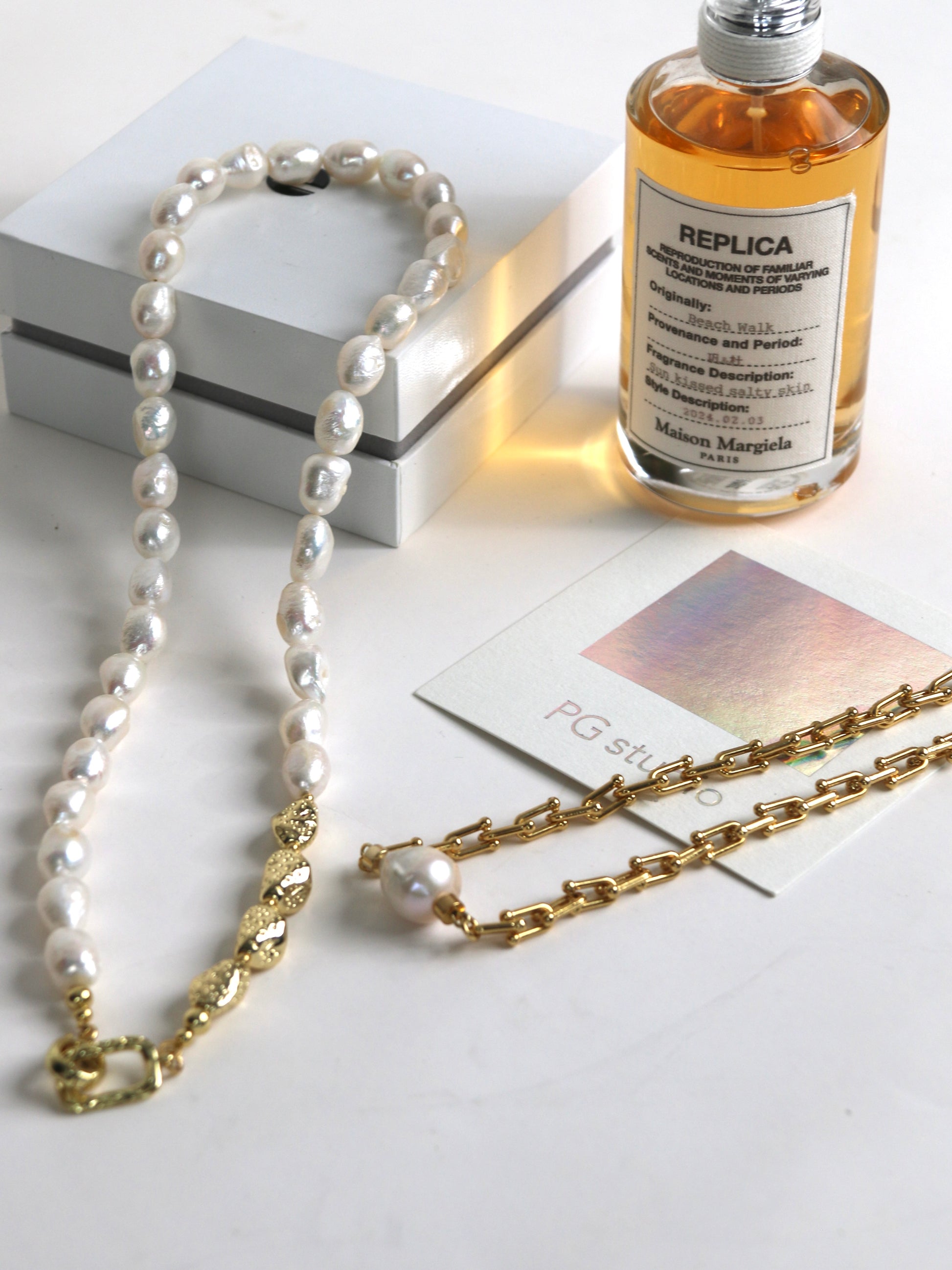 Pearl necklace for men and gold chain on a white surface with a bottle labeled 'Replica' and a book.