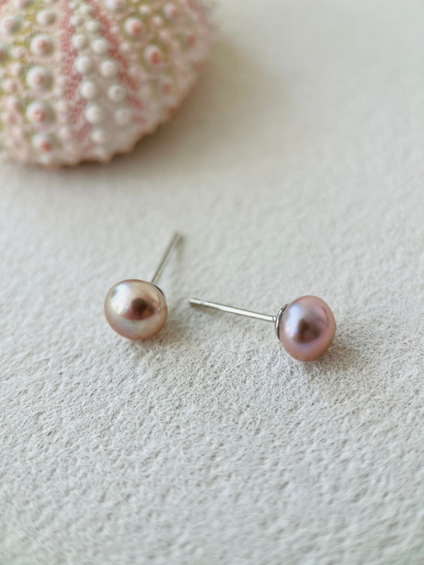 Purple Pearl stud earrings on a textured surface with a shell in the background