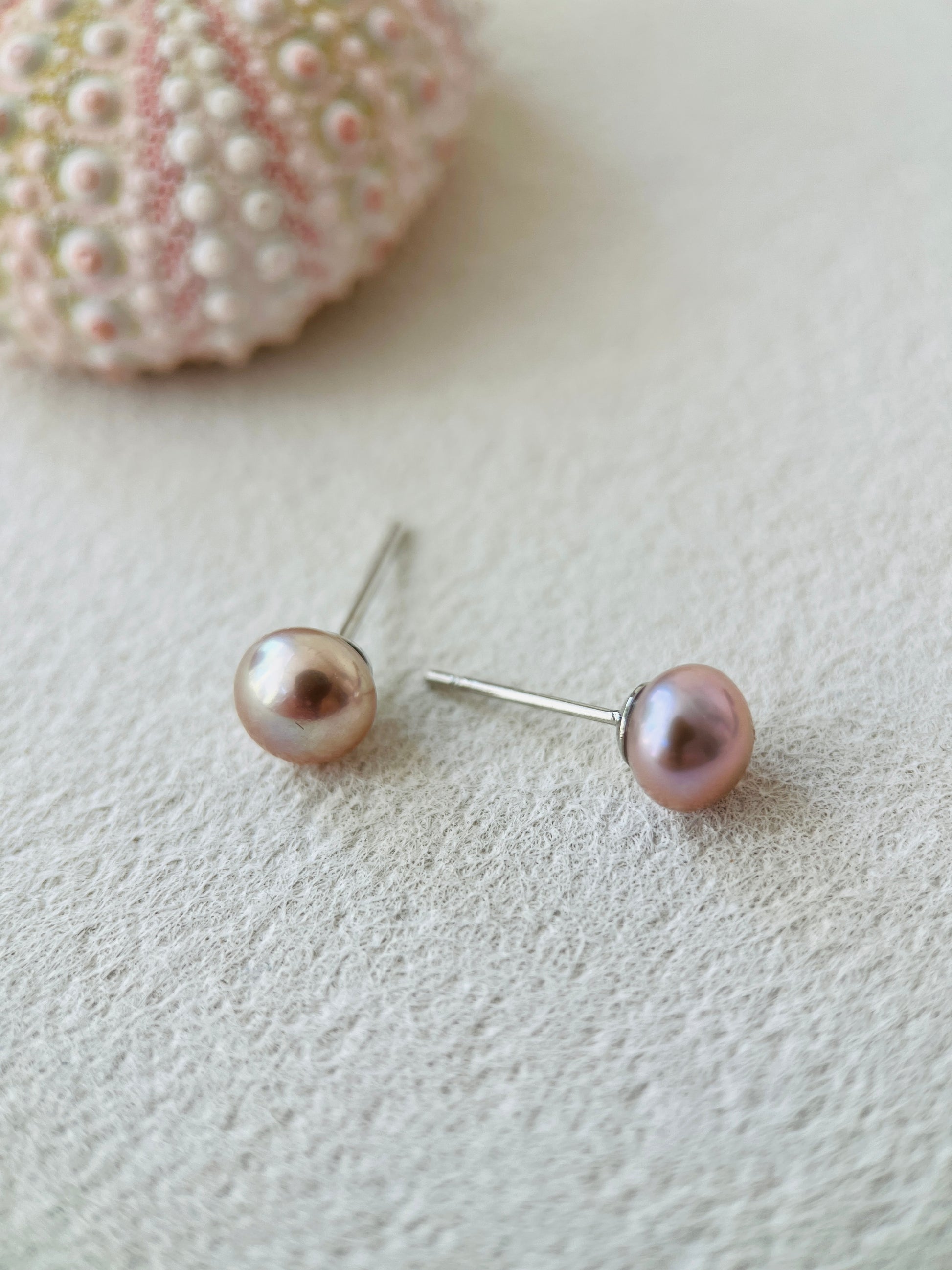 Purple Pearl stud earrings on a textured surface with a shell in the background