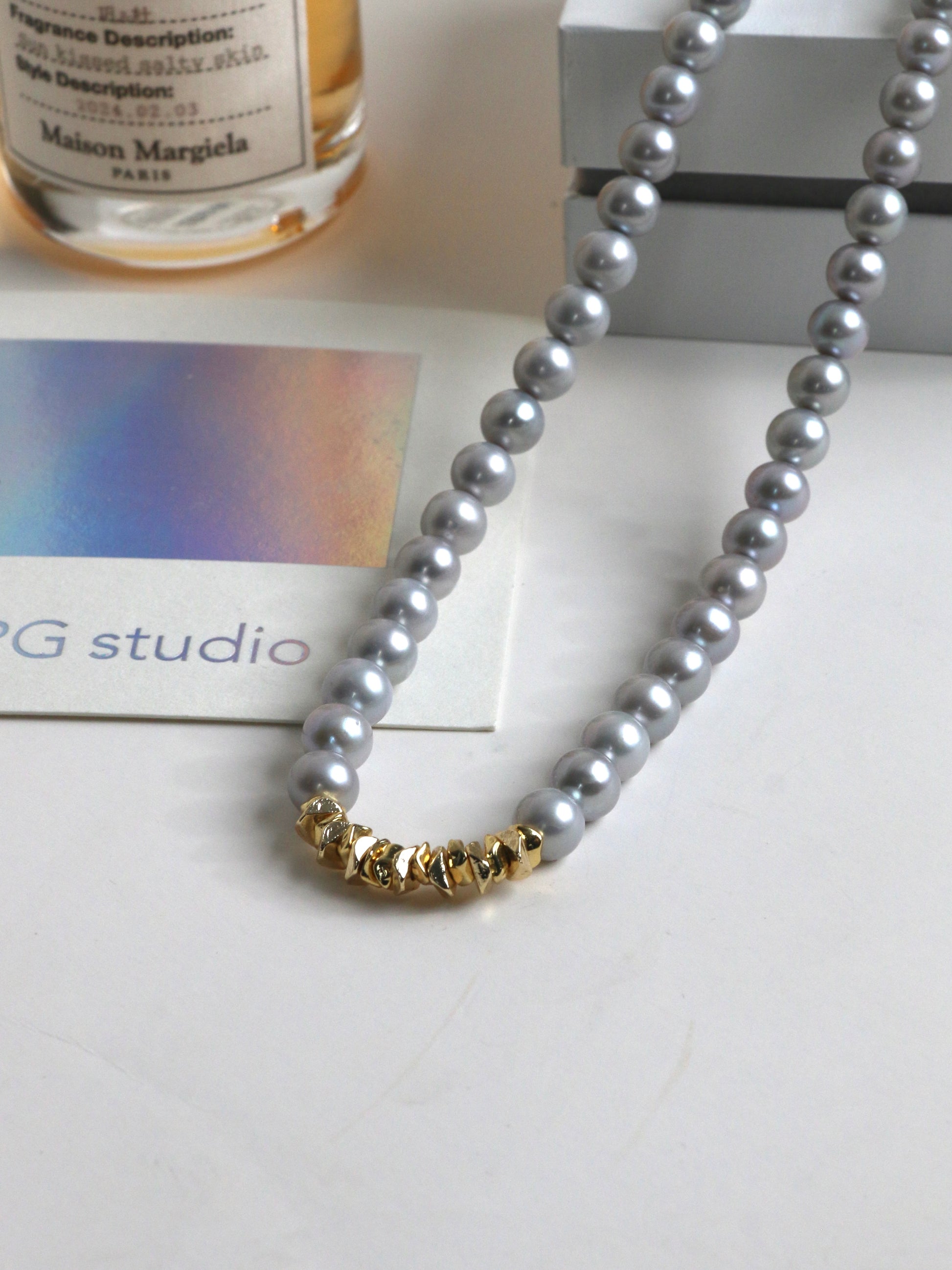Silver Pearl necklace for men with gold clasp on a white surface with a bottle and card in the background
