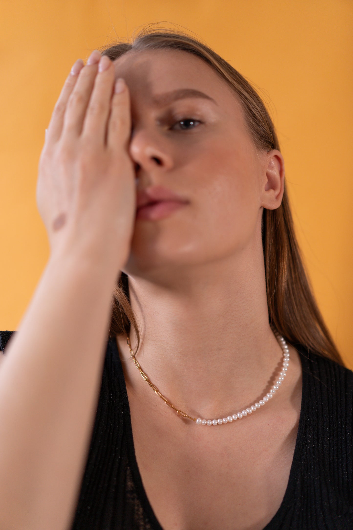 Woman wearing half pearl half chain necklace with hand on face against a yellow background 