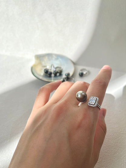 Hand wearing a ring with a large tahitian black pearl and smaller gemstones on a neutral background