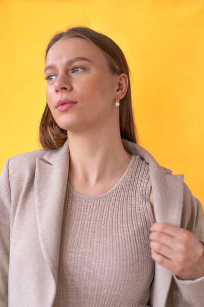Woman wearing a beige blazer and golden pearl earrings over a ribbed sweater against a yellow background