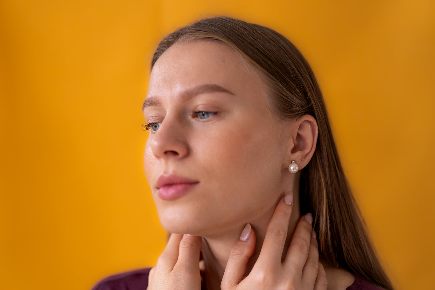 Woman touching her neck wearing pearl earrings against a mustard yellow background