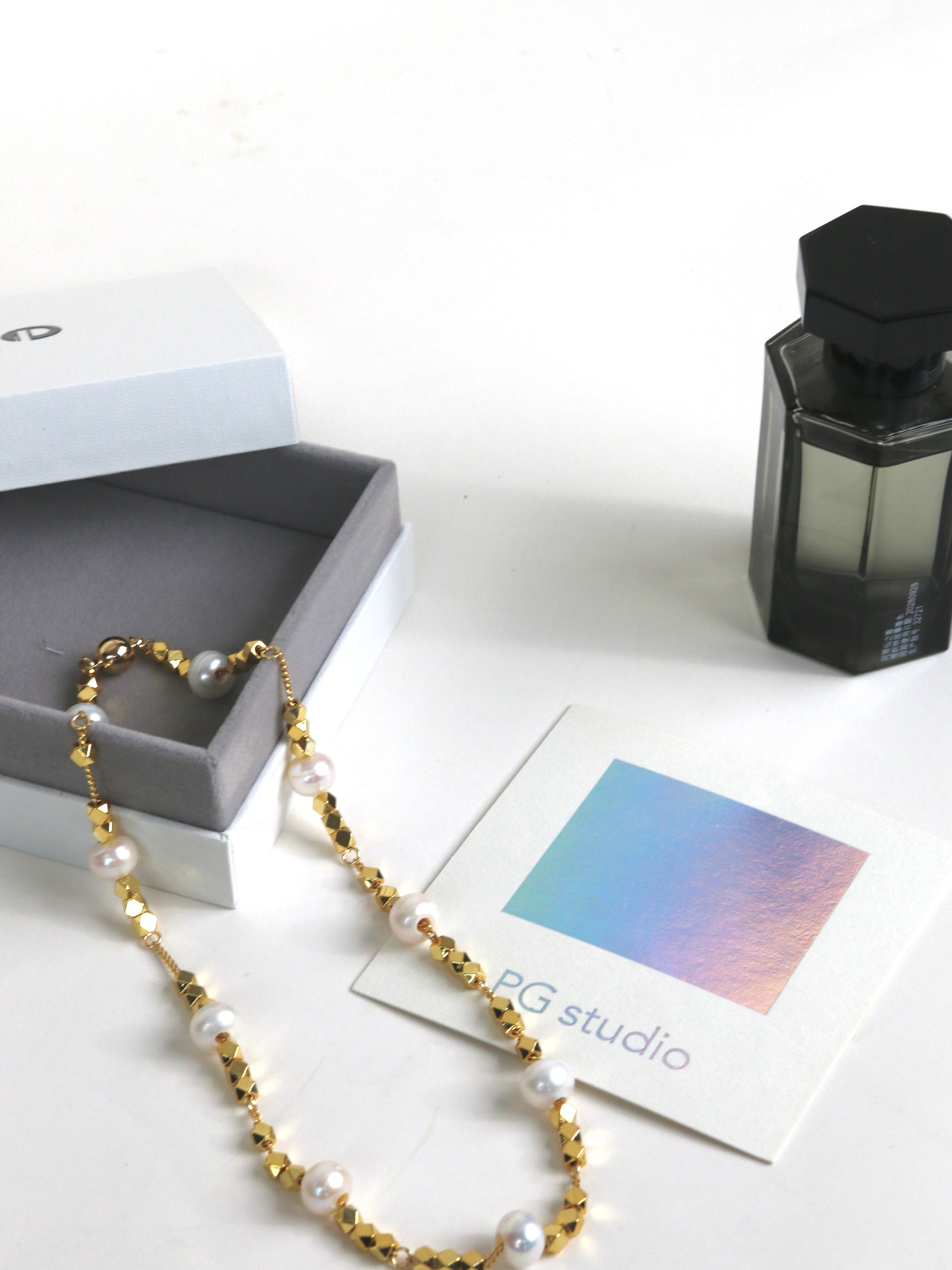 Pearl necklace for men with gold accents on a white surface with a box and perfume bottle in the background.
