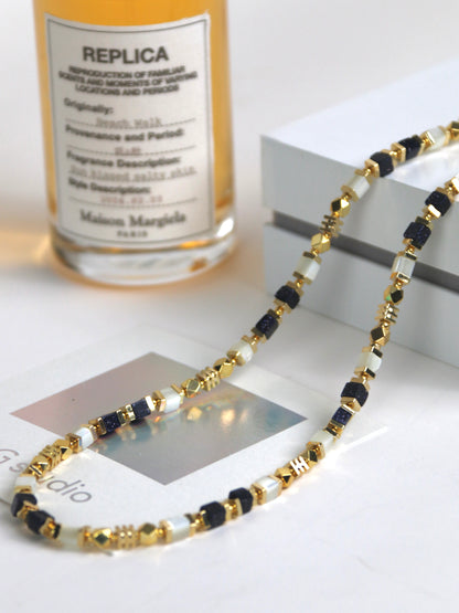 Necklace for men with gold, black, and white agate on a white surface with a perfume bottle in the background.