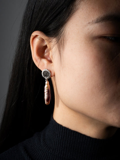 Close-up of a person wearing a dangling earring with a dark stone and pink pearl on a gray background