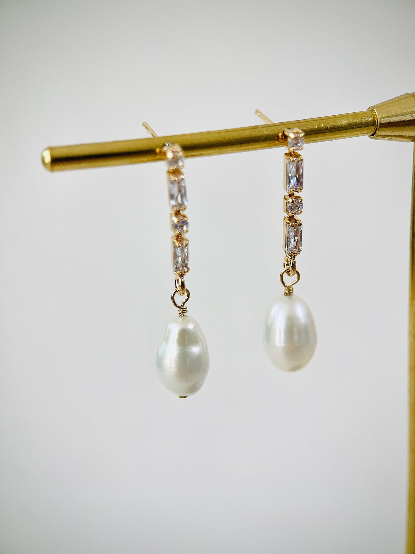 Pearl earrings with gold and clear gemstone accents on a gold stand against a light gray background