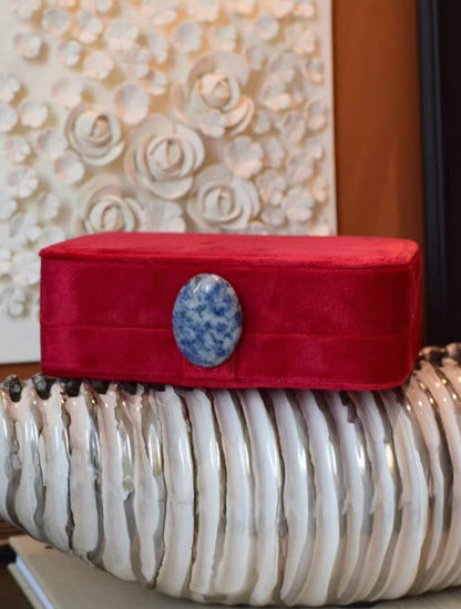 Roby red jewelry box with a blue stone on top, placed on a shell surface with floral artwork in the background.