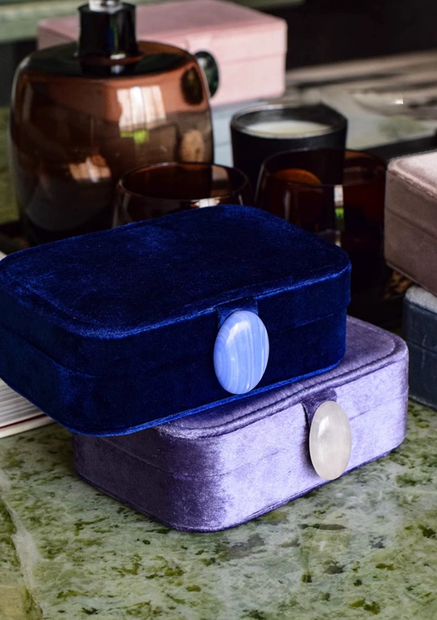 Blue and purple velvet jewelry boxes on a marble surface with a candle and perfume bottle in the background.