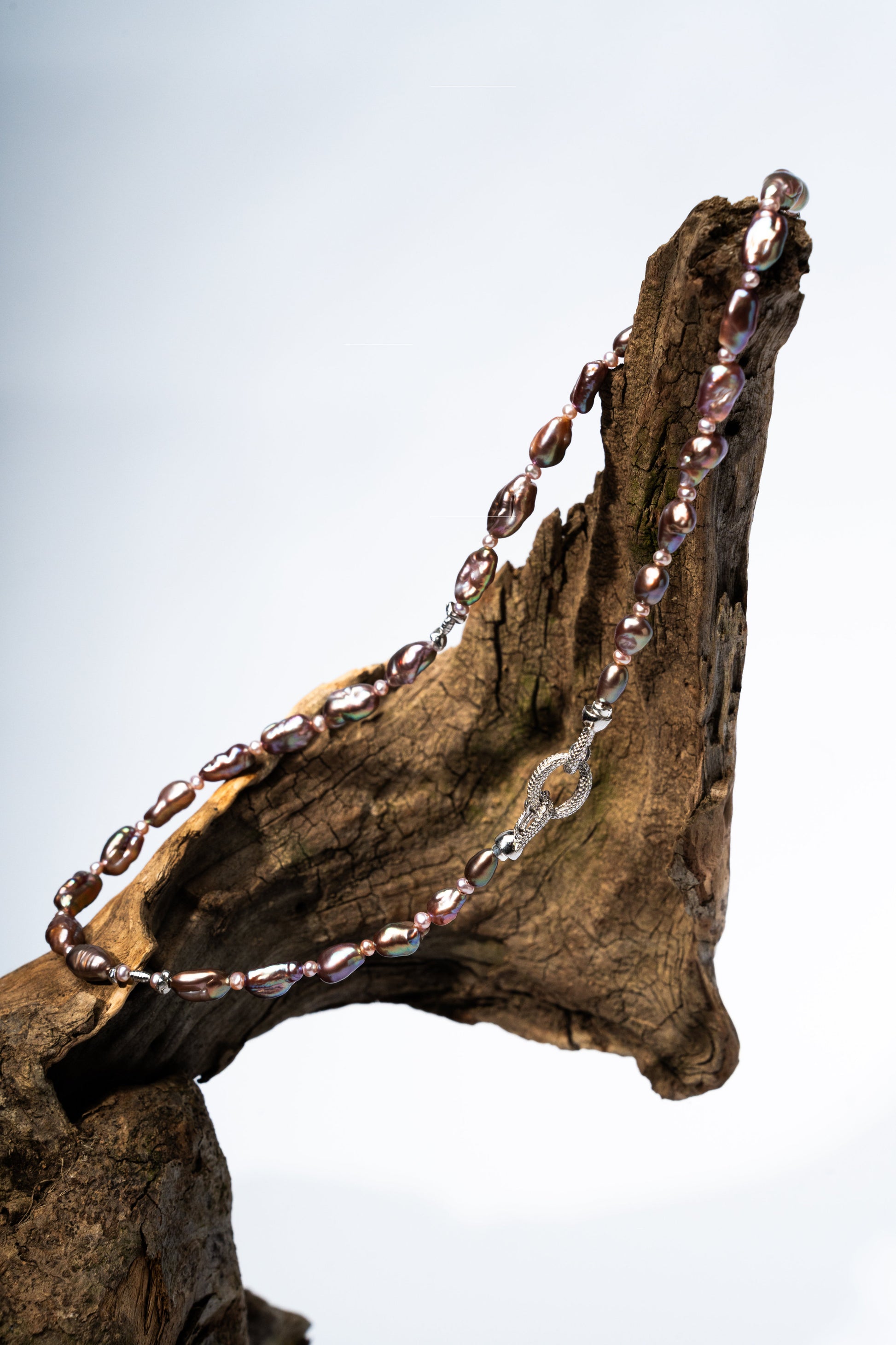 Necklace with purple Keshi pearls draped over a piece of driftwood against a light blue background
