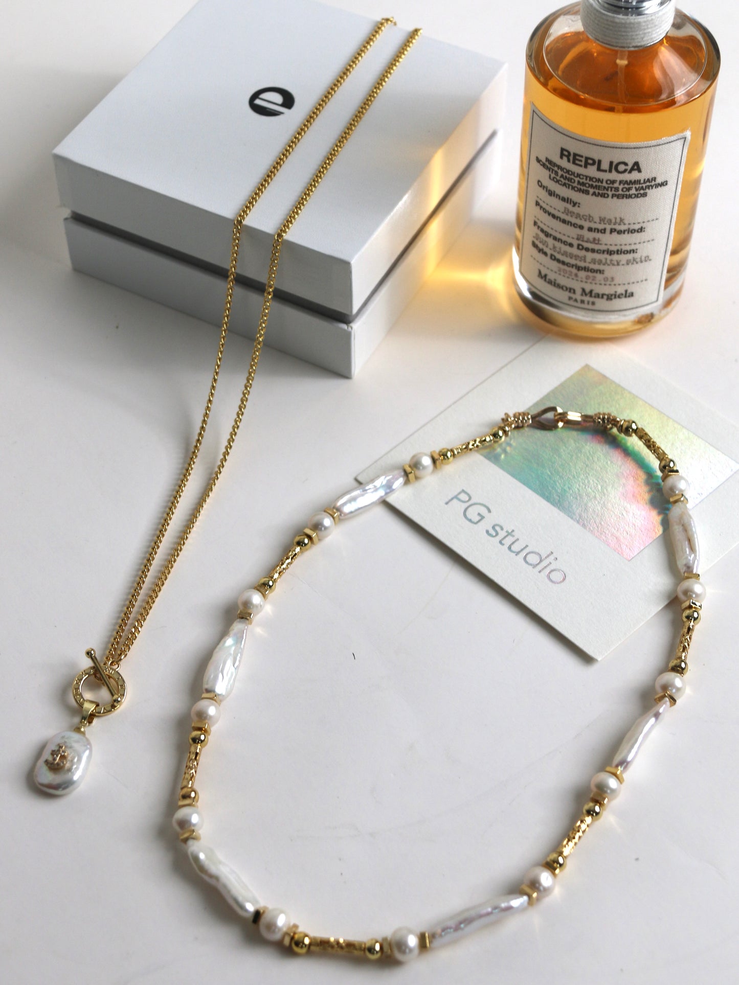Necklace with gold and pearl details on a white surface with a bottle and packaging in the background.