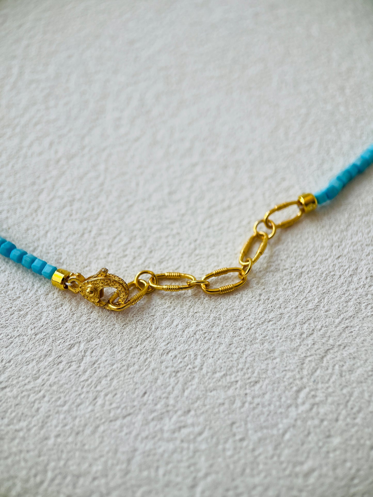 Gold chain clasp on a necklace with blue beads on a light gray background